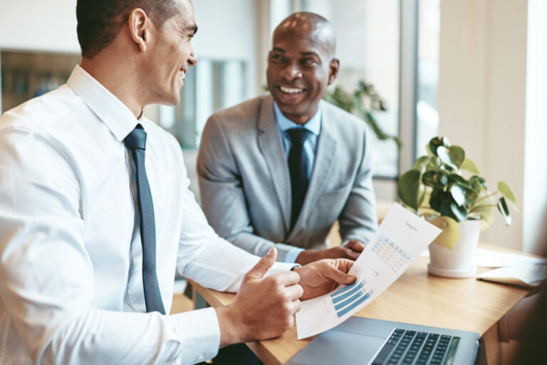 Two smiling diverse businessmen using a laptop and discussing charts and graphs on paper while sitting at a desk in an office